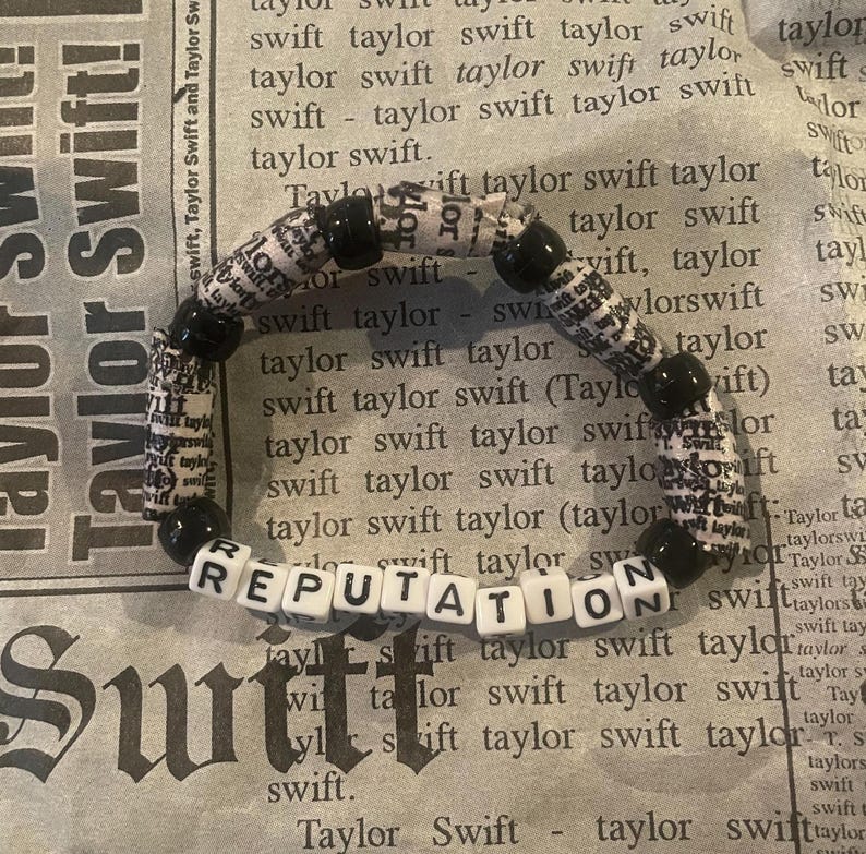 K&ouml;nnte beinhalten: Ein schwarz-wei&szlig;es Perlenarmband mit dem Wort "REPUTATION" in wei&szlig;en Perlen. Die Perlen bestehen aus Zeitungsartikeln mit dem Wort "Taylor Swift" darauf.