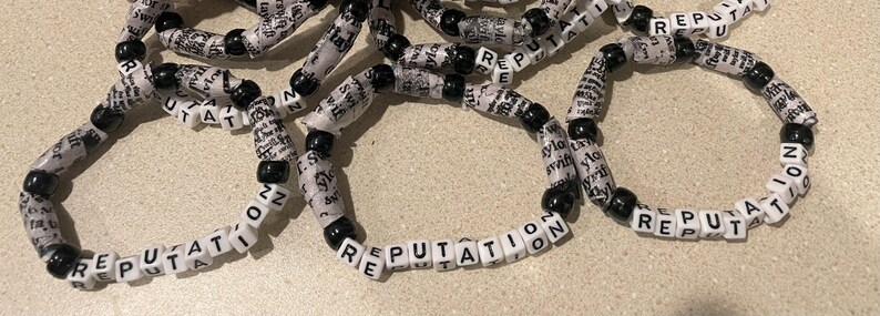 K&ouml;nnte beinhalten: Nahaufnahme von mehreren schwarzen und wei&szlig;en Perlenarmb&auml;ndern, auf denen jeweils das Wort "REPUTATION" steht.