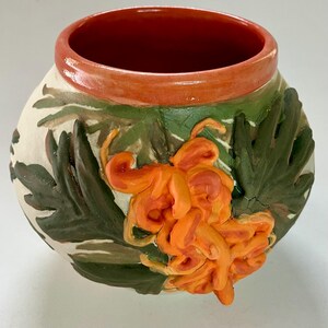 May include: A ceramic vase with a cream base, decorated with raised orange flowers and green leaves. The rim is orange, and the interior is a darker orange. The vase has a rounded shape.