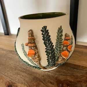 May include: A cream-coloured ceramic vase with a wide opening and a green interior. The exterior features raised, textured botanical motifs including dark green fern leaves and two brown, speckled stalks adorned with bright orange, shelf-like fungi.