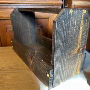 May include: A dark stained wooden tote with a handle. The rectangular box is constructed with visible wood grain and is held together with gold-colored fasteners. The tote is designed for carrying items.