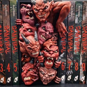 Puede incluir: Una escultura de estantería con múltiples caras de demonios rojos con cuernos y dientes afilados. La escultura está rodeada de libros titulados "Gannibal" de Masaaki Ninomiya. Los números 3 a 9 son visibles en los lomos de los libros.