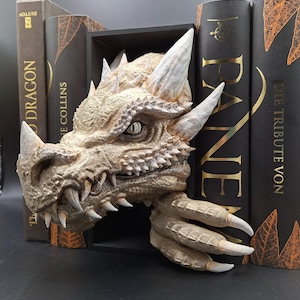 Könnte beinhalten: Ein detailliertes Drachenkopf-Buchstütze in Beige und Weiß, mit scharfen Hörnern und Zähnen. Der Drachenkopf ist zwischen mehreren dunkel gefärbten Büchern platziert und schafft ein einzigartiges Bücherregal-Accessoire.