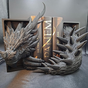 Könnte beinhalten: Ein dunkelgraues Drachen-Buchstützen-Set. Das Set enthält einen Drachenkopf und -schwanz, jeweils in einer rechteckigen Basis. Der Drachenkopf blickt nach links, und der Schwanz krümmt sich nach rechts. Die Buchstützen sind auf beiden Seiten eines Bücherstapels platziert.
