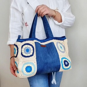 May include: A large, crocheted tote bag with denim panels. The bag features a cream-colored crochet base with blue and white circular patterns. The denim panels are a dark blue color and are sewn into the sides of the bag. The bag has two long denim handles.