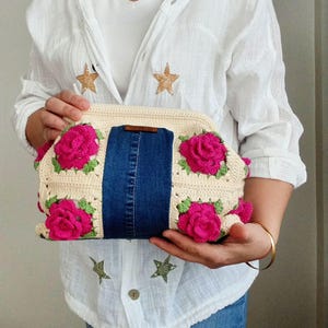 Peut inclure: Une pochette en crochet avec un panneau en jean et des accents de fleurs roses. Le sac est tenu par une personne portant une chemise blanche et un jean bleu.