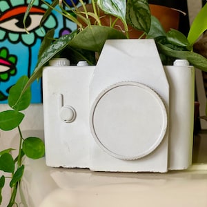 May include: White ceramic camera planter with a green plant growing out of the top. The planter is sitting on a white surface.