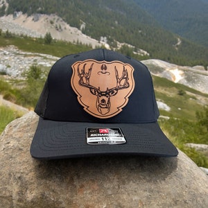 May include: Black trucker hat with a brown leather patch featuring a deer head design. The hat has a curved brim and a mesh back. The patch has a shield shape with the deer's antlers prominently displayed. The hat is adjustable.