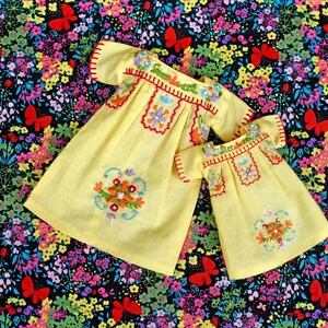 May include: Two yellow dresses with short sleeves, featuring colorful floral embroidery around the neckline and a matching floral motif on the lower front. The embroidery includes red, orange, blue, and green thread with small sequins.