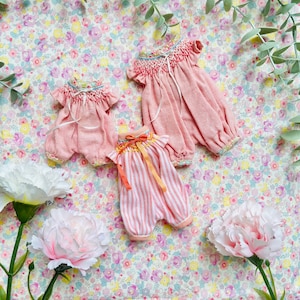May include: Three infant rompers featuring smocked necklines and ruffled sleeves. Two rompers are solid dusty pink with floral trim, and one features pink and white vertical stripes with an orange ribbon tie at the center of the neckline.