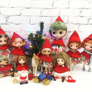 May include: A collection of dolls dressed in red hooded capes with white polka dots and floral dresses, arranged around a small artificial Christmas tree with miniature gifts, a small Santa Claus figure, a deer figurine, and a rabbit figurine.