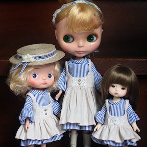 May include: Three dolls wearing matching blue and white striped dresses with beige pinafores. The left doll has blonde hair and a straw hat. The center doll has blonde hair in a bun and grey boots. The right doll has brown hair and brown shoes.