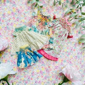 May include: Four small dresses featuring intricate smocking at the necklines. One dress is cream with gold dots, one has a floral print with yellow smocking, one is a blue floral print, and one is a pink floral print with a pink hem and lace trim.