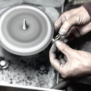 Könnte beinhalten: Nahaufnahme einer Hand, die einen Ring hält, der auf einem rotierenden Polierrad poliert wird. Der Ring wird von einer Hand mit schmutzigen Fingern gehalten. Das Polierrad dreht sich schnell und erzeugt einen Unschärfe-Effekt.