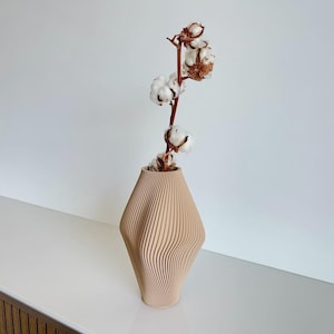 Puede incluir: Un jarrón de madera beige con un diseño geométrico único. El jarrón tiene líneas verticales y forma de diamante. Contiene un tallo de algodón con bolas de algodón blancas y tallos marrones. El jarrón está sobre una superficie blanca.
