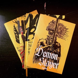 May include: Three comic books stacked on top of each other. The top book is yellow with black text that reads "Demon Slayer". The middle book is yellow with black text that reads "Text and Graphic Design by Pelle F. Johan and Dean...