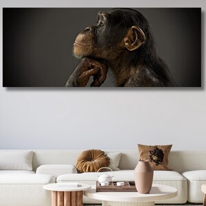 May include: A rectangular canvas print featuring a chimpanzee in profile, set against a dark gray background. The chimpanzee has brown fur and is resting its chin on its hand, appearing thoughtful. The artwork is displayed above a white sofa.
