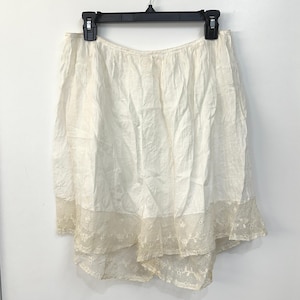 May include: An off-white skirt featuring a gathered waist and a textured fabric finish. The hem is adorned with a wide, intricate lace trim that includes floral embroidery and a delicate mesh pattern, creating a layered and detailed appearance.