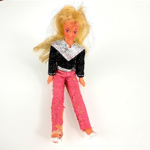 May include: A vintage doll with blonde hair, wearing a black and silver top, pink pants, and white shoes. The doll's outfit has a textured appearance. The doll is posed on a white surface.