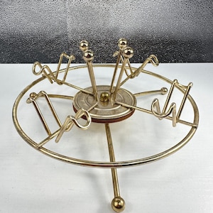 May include: A gold-toned metal carousel-style stand featuring a circular outer ring, a central rotating disc, and multiple upward-curving hooks designed for hanging items. The structure includes a central vertical post with a rounded finial top.