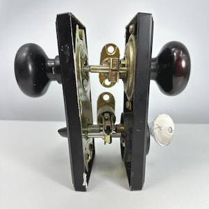 Peut inclure: Un ensemble de serrure de porte vintage composé de deux boutons sphériques marron foncé fixés sur des plaques métalliques rectangulaires noires. Le mécanisme interne est visible, avec des pièces en laiton, un loquet et une clé argentée.