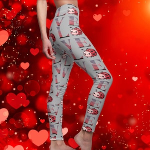 May include: Gray leggings with a repeating pattern of red ladybugs and the word "LOVE" in white. The leggings are on a model against a red background with hearts.