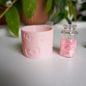 Puede incluir: Un recipiente cilíndrico rosa claro con diseños de lazos en relieve, junto a una pequeña botella de vidrio llena de purpurina rosa. Los objetos están sobre una superficie blanca, con follaje verde en el fondo.