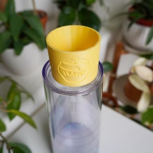 Puede incluir: Un manguito de vaso de plástico amarillo con un diseño de tarro de miel y la palabra "HONEY" en relieve. El manguito está colocado sobre un vaso transparente iridiscente, con plantas verdes en el fondo.