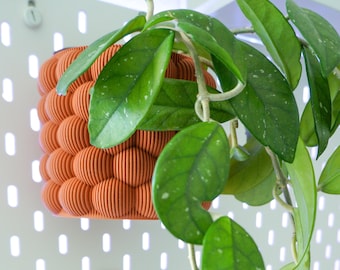 May include: An orange, textured, 3D-printed plant pot with a spherical design, mounted on a white pegboard. The pot holds a green plant with large, speckled leaves. The pot's design is made of many vertical, parallel lines.