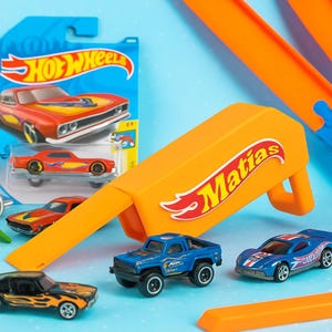 Può includere: Un assortimento di macchinine e pezzi di pista Hot Wheels su sfondo azzurro. Il set include un pick-up blu, un'auto da corsa blu e rossa e un'auto nera con fiamme. Un pezzo di pista arancione è etichettato "Matias".