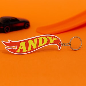 Puede incluir: Un llavero personalizado con forma de llama roja, borde blanco y el nombre ANDY escrito en letras mayúsculas amarillas. El llavero está unido a una anilla de metal plateado mediante una pequeña cadena metálica.
