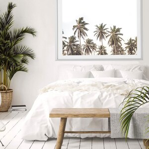 May include: A white bed with white bedding and a wooden bench in front of it. A framed print of palm trees is hanging on the wall behind the bed.