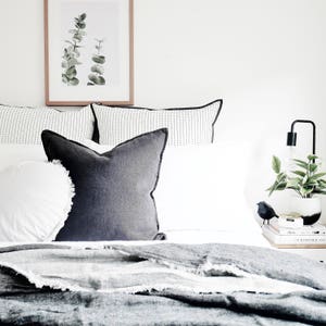 May include: A white bed with a black and white patterned pillow, a dark gray pillow, and a white blanket. A framed print of green leaves hangs above the bed. A black lamp with a white shade sits on a nightstand next to the bed.