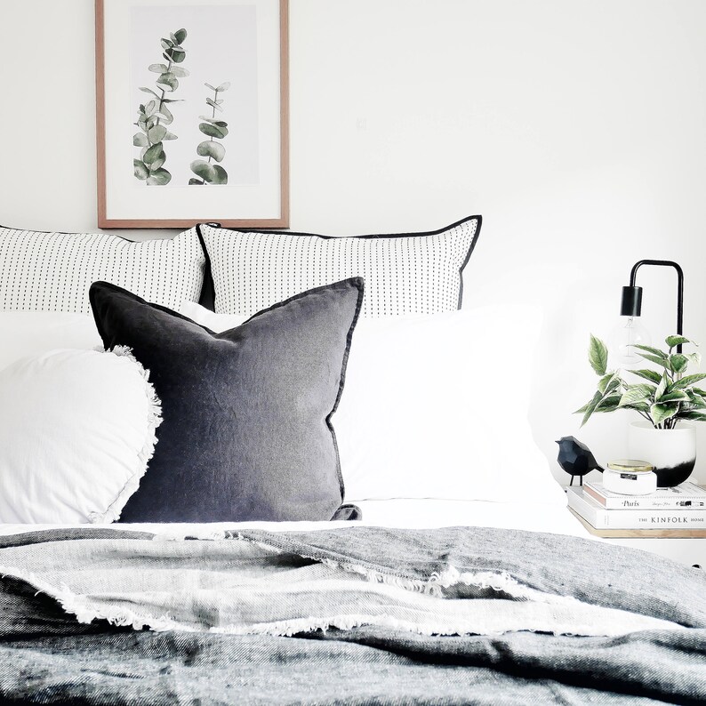 May include: A white bed with a black and white patterned pillow, a dark gray pillow, and a white blanket. A framed print of green leaves hangs above the bed. A black lamp with a white shade sits on a nightstand next to the bed.