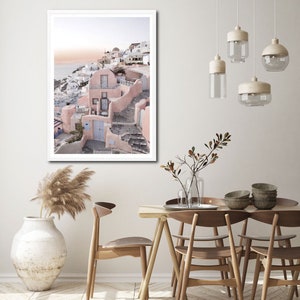 May include: A framed photograph of a pink and white building with a blue door, set against a backdrop of a blue ocean and a cloudy sky. The building is situated on a hillside with steps leading up to it. The photograph is displayed on a white wall in a dining room setting with a wooden table and chairs.