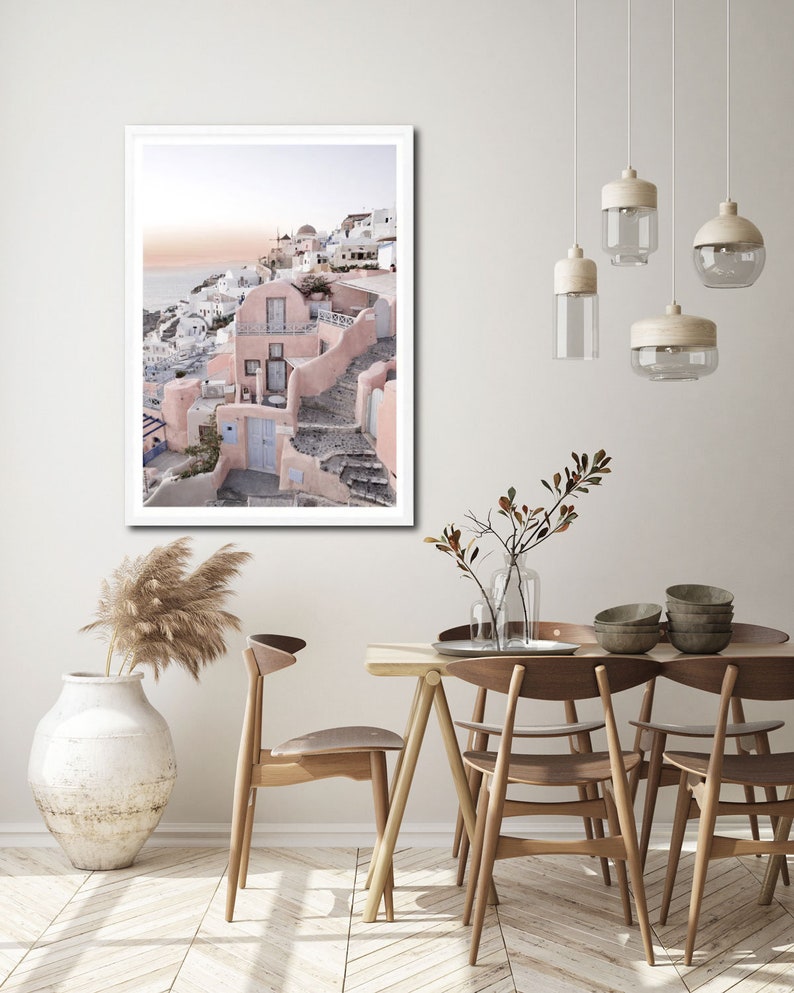 May include: A framed photograph of a pink and white building with a blue door, set against a backdrop of a blue ocean and a cloudy sky. The building is situated on a hillside with steps leading up to it. The photograph is displayed on a white wall in a dining room setting with a wooden table and chairs.