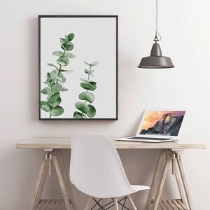 May include: A framed watercolor print of two green eucalyptus branches on a white background. The print is hanging on a white wall above a wooden desk with a white chair and a laptop.