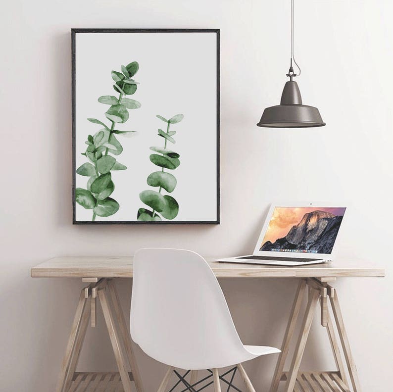 May include: A framed watercolor print of two green eucalyptus branches on a white background. The print is hanging on a white wall above a wooden desk with a white chair and a laptop.