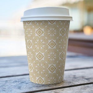 May include: A disposable coffee cup with a white lid. The cup features a repeating gold geometric pattern on a cream background. The cup is sitting on a wooden surface.