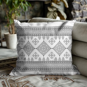 May include: A white decorative pillow with a gray geometric pattern. The pattern includes diamond, swirl, and linear designs. The pillow is on a beige sofa and patterned blanket.