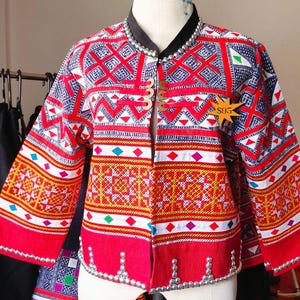 Pode incluir: Uma jaqueta vibrante e estampada com um design geométrico vermelho, branco e azul. A jaqueta apresenta uma gola preta, botões dourados decorativos e enfeites prateados nas bordas. A jaqueta é exibida em um manequim.