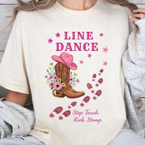 May include: Cream-colored t-shirt with a graphic featuring a cowboy boot, pink hat, and floral accents. The text "LINE DANCE" is above the design, with star accents. Below, the text reads "Step. Touch. Kick. Stomp."