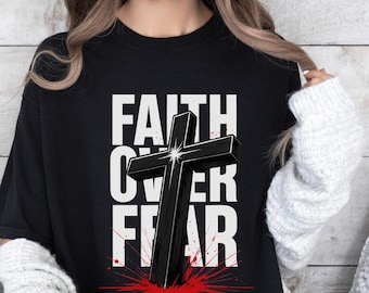Op de afbeelding: Zwart T-shirt met de witte tekst "FAITH OVER FEAR" en een 3D zwarte kruisafbeelding. Het kruis staat tegen een rood spatterontwerp, wat een opvallende visuele impact creëert.