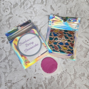 Może przedstawiać: Dwa małe, tęczowe woreczki, w jednym z nich znajduje się różowa masa do zabawy, a w drugim nadruk z dinozaurami. Woreczek z masą do zabawy ma etykietę z napisem "Brontosaurus Berry Cheesecake". Woreczek z nadrukiem dinozaurów ma etykietę z napisem "Tiny Dino Creations".