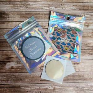 May include: Two small, iridescent, zippered bags with labels. The bag on the left has a white label with the text "Oatmeal Bread Omnivore". The bag on the right has a label with a dinosaur pattern and the text "Tiny Dino Creations".