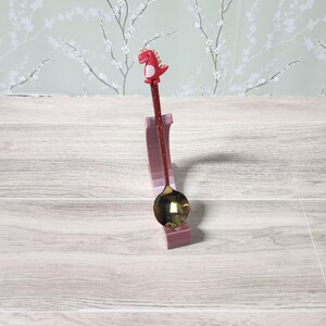 May include: A decorative spoon featuring a red dinosaur figure atop a glittery red handle. The spoon has a gold-coloured bowl and is displayed on a pink stand. The background features a light green floral wallpaper.
