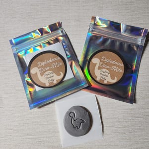 Może przedstawiać: Dwie małe srebrne torebki z zamkami błyskawicznymi, każda zawierająca małe brązowe kółko z tekstem "Diplodocus Dino-Mite Tiny Dry Creation Pretty" i sylwetkę szarego dinozaura. Oddzielna szara plastelina w kształcie dinozaura jest pokazana na białym tle.