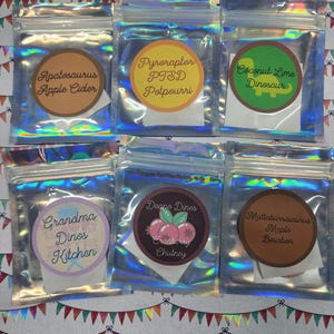 Może przedstawiać: Sześć małych, przezroczystych, zamykanych torebek z kolorowymi etykietami. Etykiety zawierają teksty takie jak "Apatosaurus Apple Cider" i "Coconut Lime Dinosaur". Torebki są ułożone na świątecznym tle z girlandami chorągiewek.