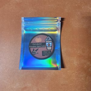 以下が含まれることがあります： ジッパー開閉式のホログラフィックポーチ。円形のラベルにはカボチャとコーヒーカップのイラストが描かれています。ラベルには「Pterodactyl Pumpkin Spice Latte」と書かれており、香りのノートが記載されています。