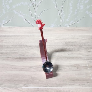 May include: A decorative spoon featuring a red dinosaur figurine. The handle is glittery red, and the bowl is silver. It rests in a pink stand on a light wood surface.